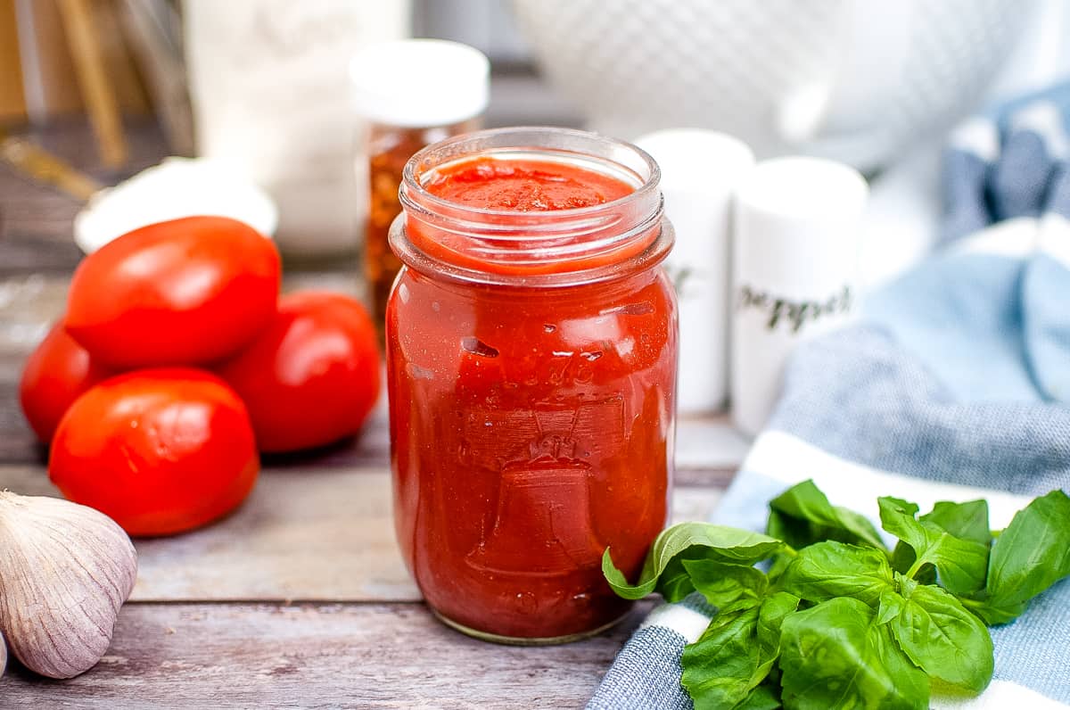 homemade pizza sauce in glass mason jar with tomatoes, salt and pepper, and basil in the background