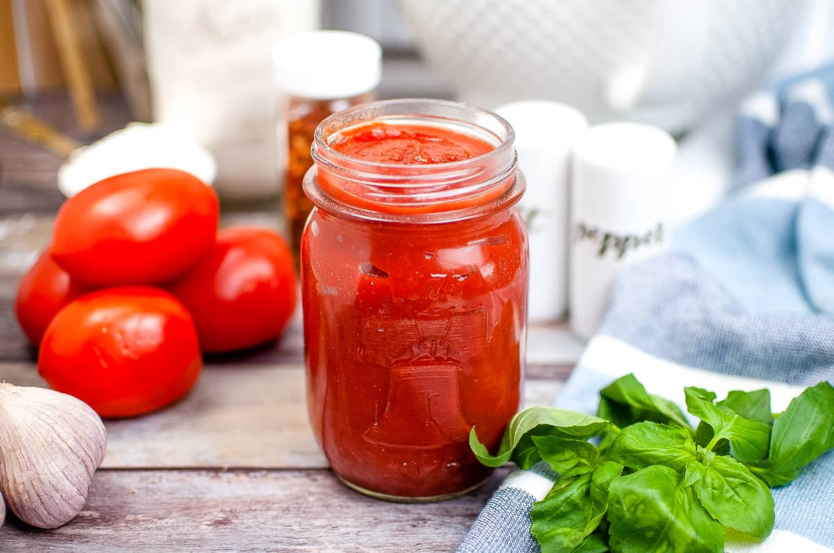 Easy homemade pizza sauce made from tomato paste in a glass mason jar with tomatoes, garlic, salt and pepper, and basil in the background.