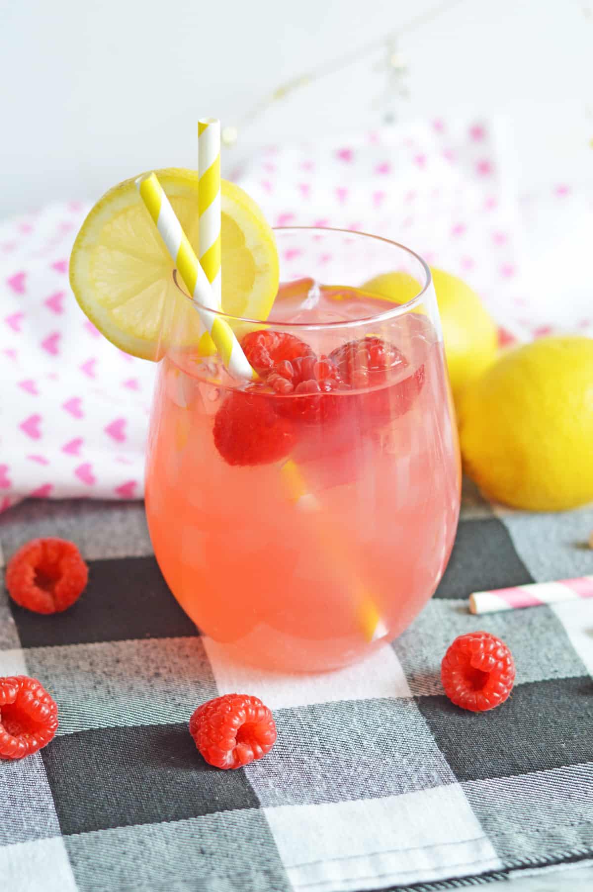 Raspberry vodka lemonade cocktail in stemless wine glass garnished with fresh raspberries and a lemon slice. Two yellow and white striped paper straws are in the glass and additional fresh raspberries are surrounding the glass.