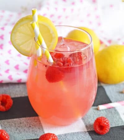 Raspberry vodka lemonade cocktail in stemless wine glass garnished with fresh raspberries and a lemon slice. Two yellow and white striped paper straws are in the glass and additional fresh raspberries are surrounding the glass.