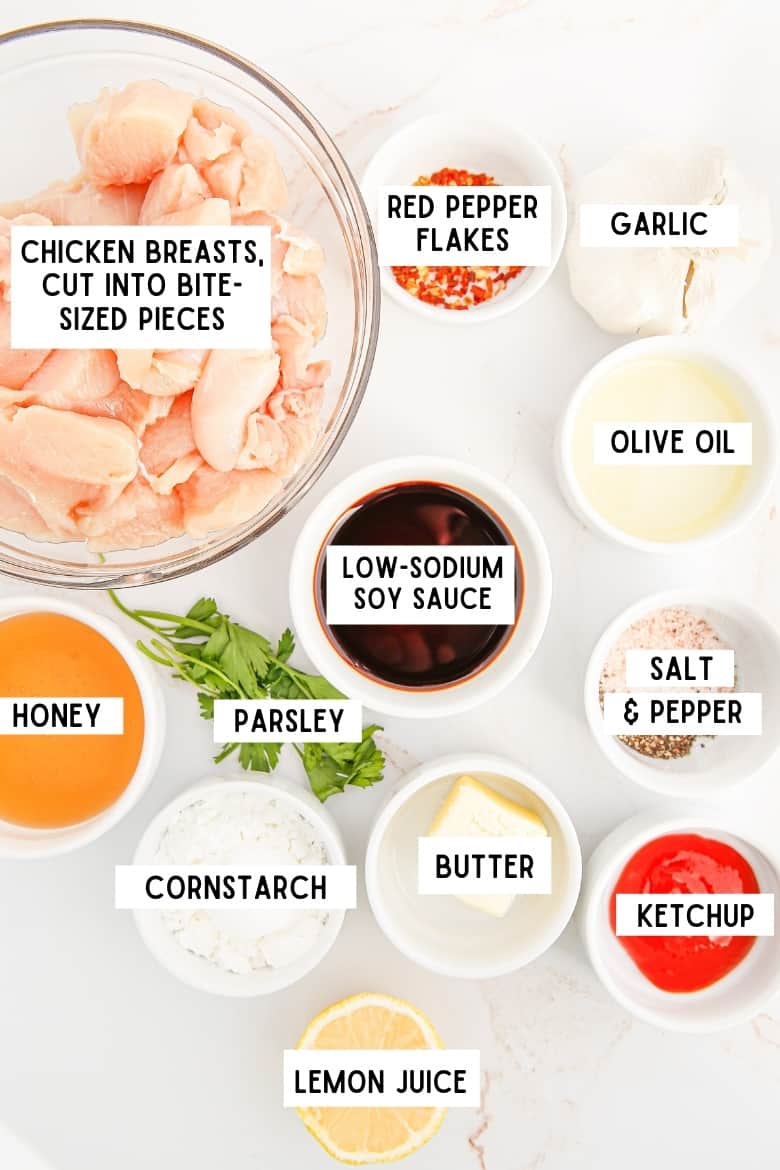ingredients in bowls on countertop: chicken breasts cut into bite-sized pieces, red pepper flakes, olive oil, garlic, soy sauce, honey, cornstarch, butter, ketchup, lemon juice, salt and pepper, and parsley