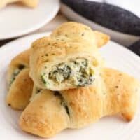 Three spinach artichoke crescent rolls on a plate, one with a bite taken out of it to show the creamy and cheesy spinach dip inside of it.