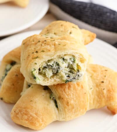 Three spinach artichoke crescent rolls on a plate, one with a bite taken out of it to show the creamy and cheesy spinach dip inside of it.