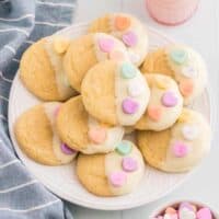 Vanilla cookies dipping in white chocolate and topped with 3 conversation heart candies. The Valentines cookies are served stacked on a white plate with a glass of milk, linen, and bowl of conversation hearts around them.