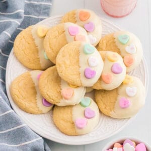 Vanilla cookies dipping in white chocolate and topped with 3 conversation heart candies. The Valentines cookies are served stacked on a white plate with a glass of milk, linen, and bowl of conversation hearts around them.