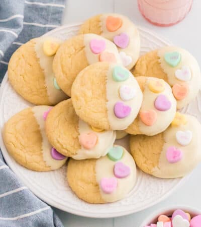 Vanilla cookies dipping in white chocolate and topped with 3 conversation heart candies. The Valentines cookies are served stacked on a white plate with a glass of milk, linen, and bowl of conversation hearts around them.