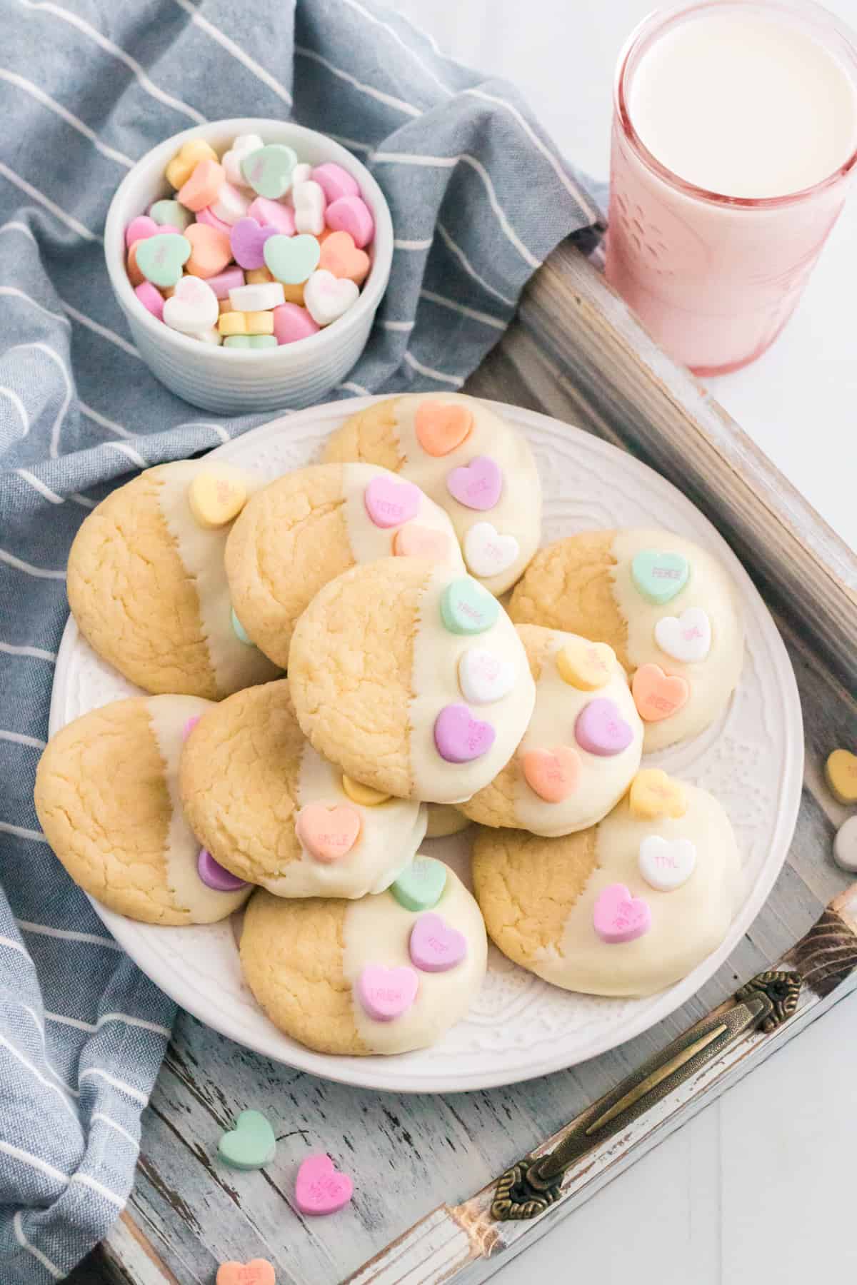 Valentines cake mix cookies dipped in white chocolate and topped with candy hearts. Glass of milk and bowl of conversation heart candies are beside the plate of cookies.