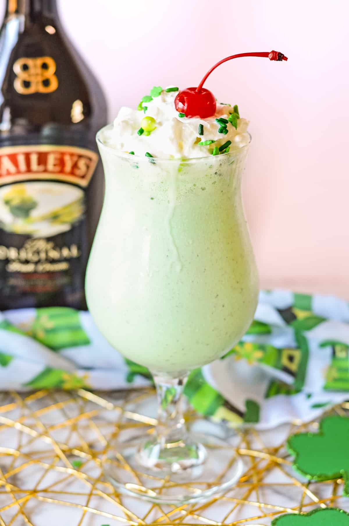 Mint green milkshake topped with whipped cream, green sprinkles, and a cherry. A bottle of baileys in background.