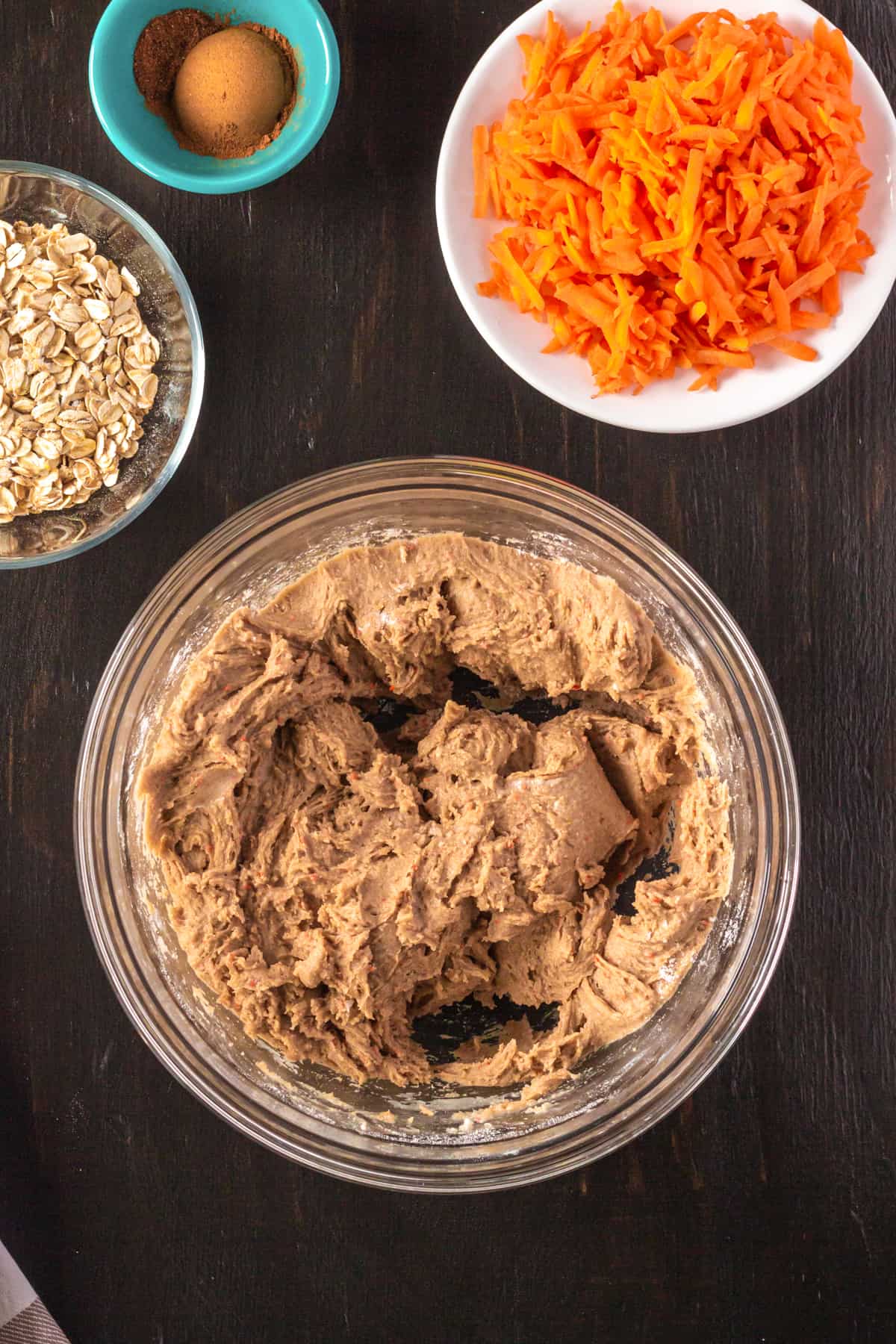 Cookie dough in mixing bowl with shredded carrots, oats, and spices nearby.