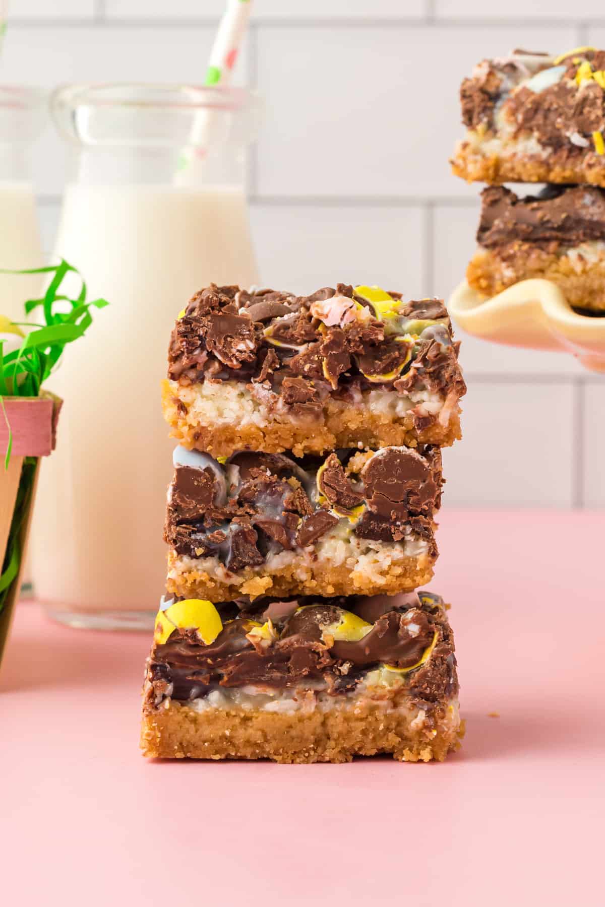 Three thick easter dessert bars stacked on top of one another with glass of milk and additional bars in the background.