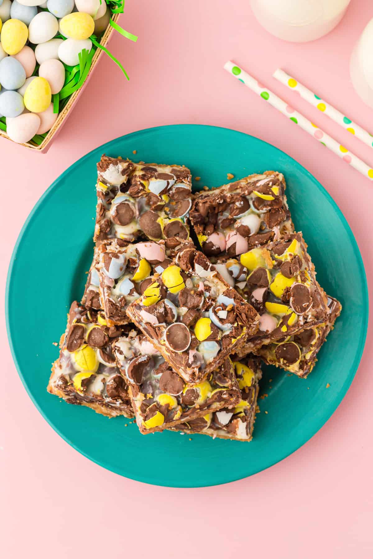 Top-down photo of mini egg cookie bars just into squares and laid on teal plate. Mini chocolate eggs, paper straws, and glasses of milk are in background.