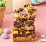 Easter dessert bars with sweetened condensed milk, chocolate chips, coconut, and chocolate eggs stacked on top of one another with top bar missing a bite.