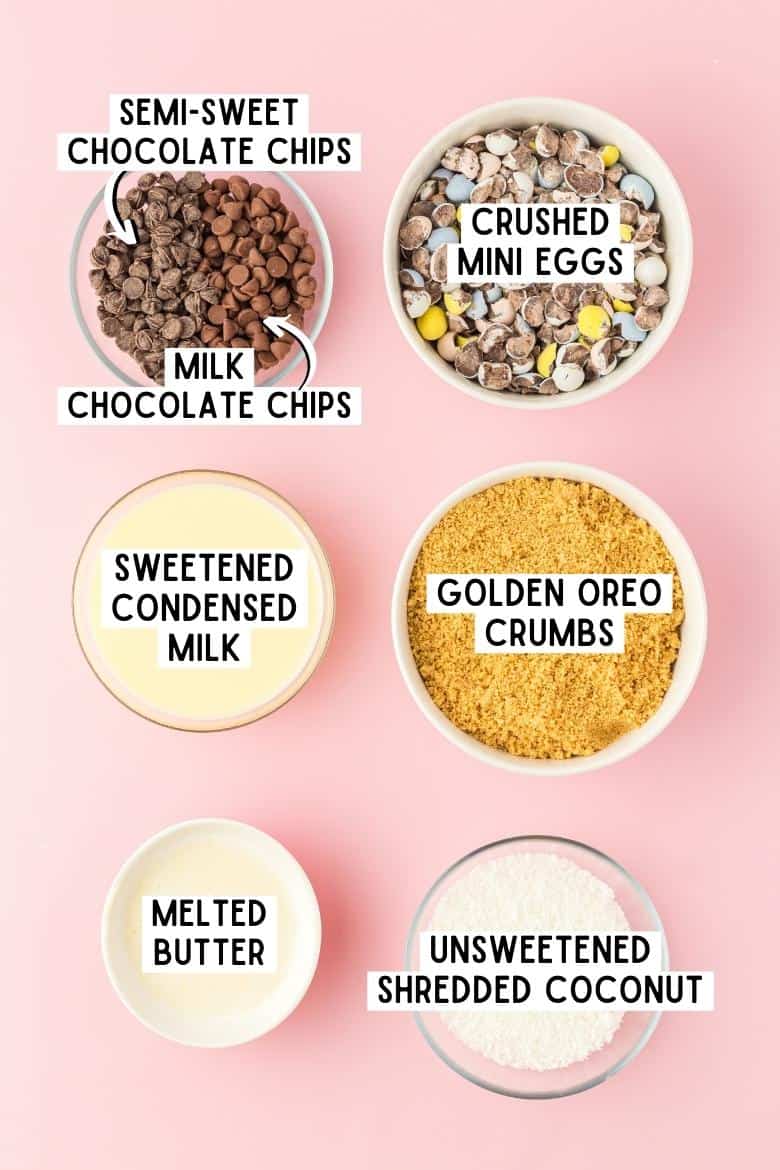 Milk chocolate chips, semi-sweet chocolate chips, crushed mini eggs, golden oreo crumbs, sweetened condensed milk, melted butter. unsweetened shredded coconut in bowls on pink surface.
