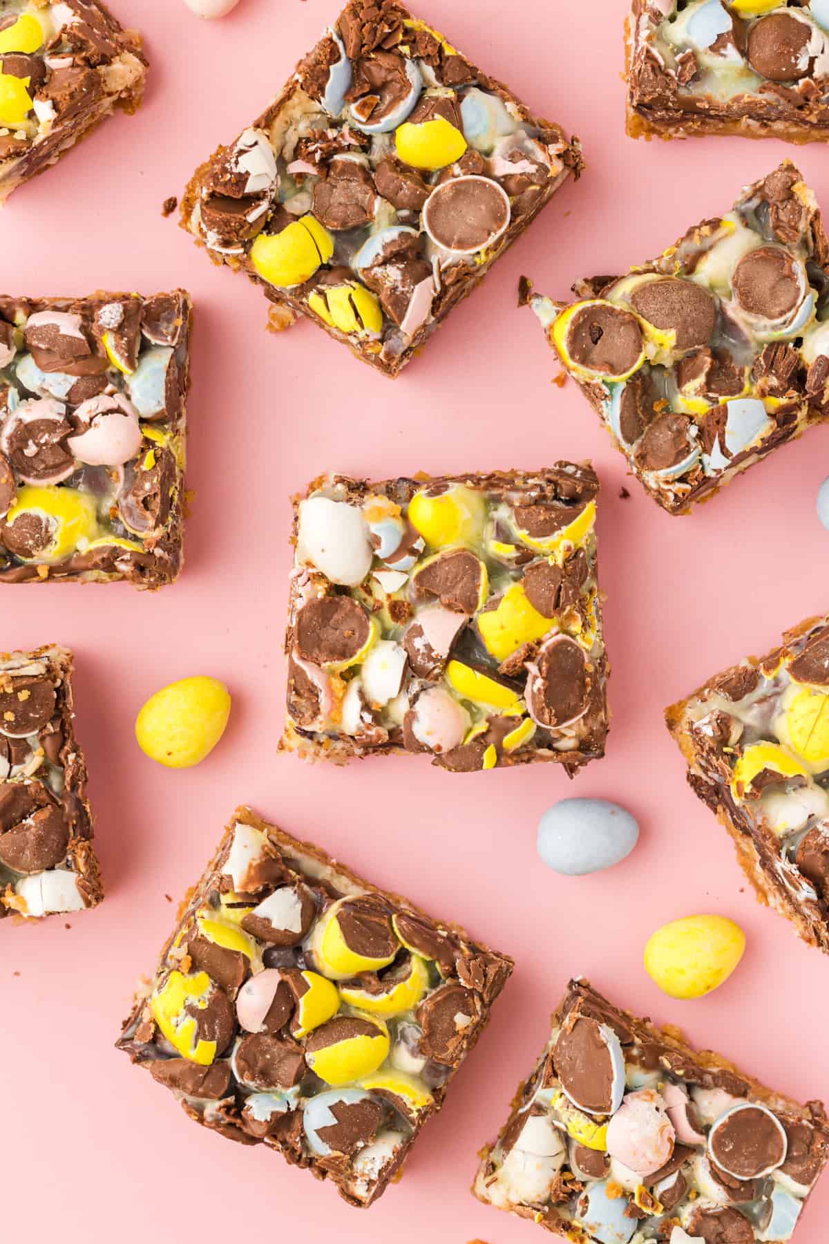 Easter magic cookie bars topped with crushed pastel mini eggs cut into squares and laid on a pink surface with additional mini eggs scattered around.