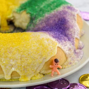King cake with glaze and green, yellow, and purple sanding sugars. A small toy baby is leaning on the cake.