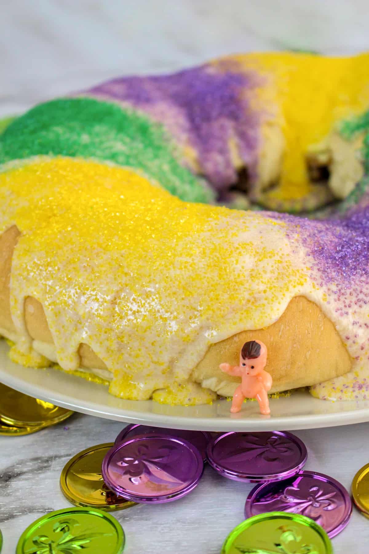 Easy homemade king cake with vanilla glaze and decorated with purple, green, and yellow sanding sugar. A small plastic baby in one the white platter next to the cake.