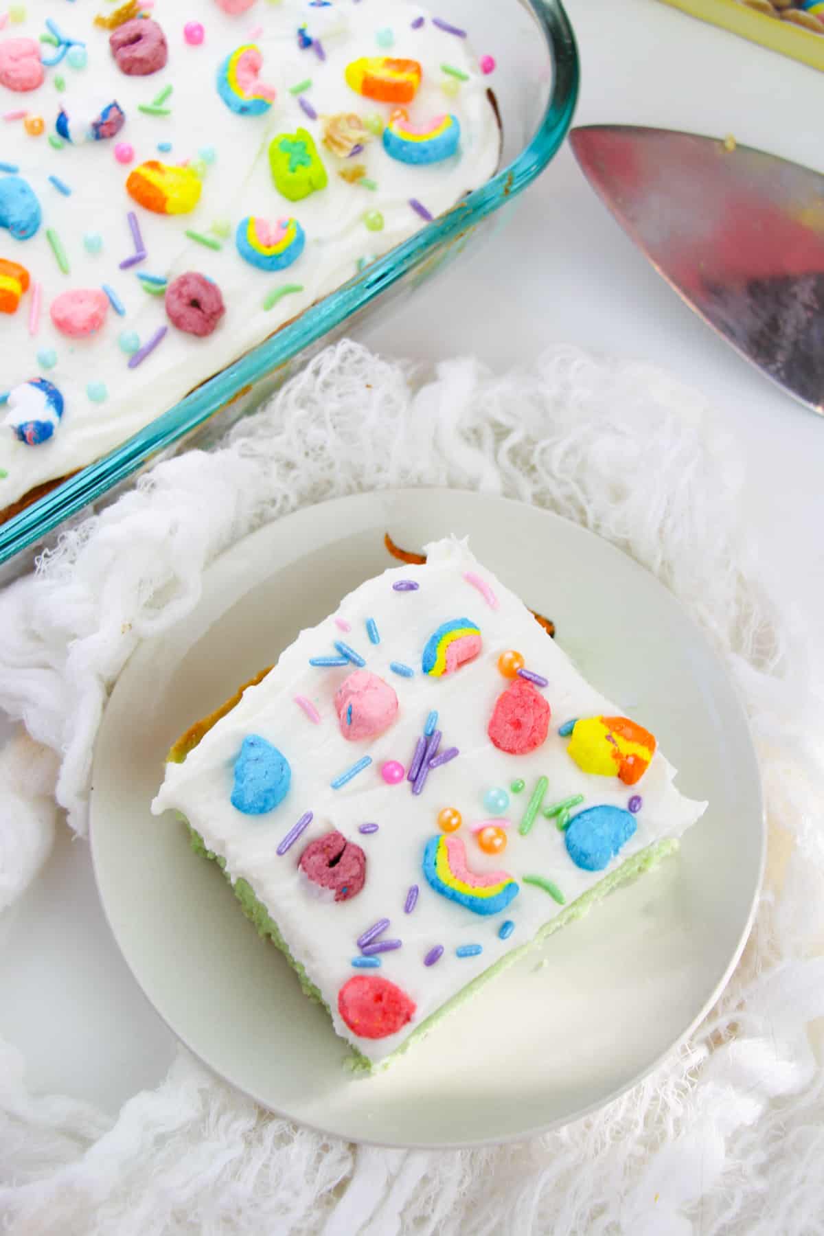 Slice of lucky charms sheet cake with white frosting topped with rainbow sprinkles and lucky charms marshmallows. Whole cake in baking pan can be seen next to the slice.