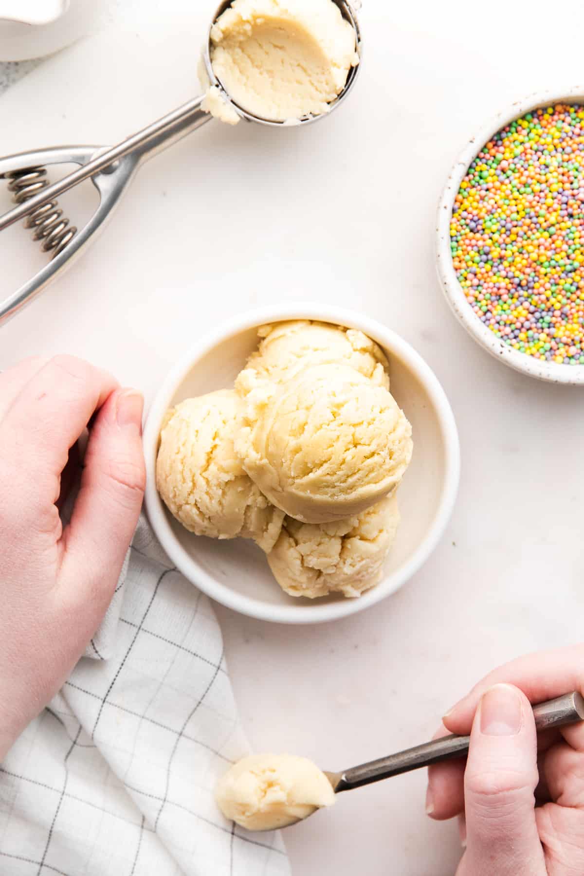 bowl of cookie sugar dough, bowl of sprinkles, cookie scoop filled with cookie dough, and hand holding spoon of cookie dough.