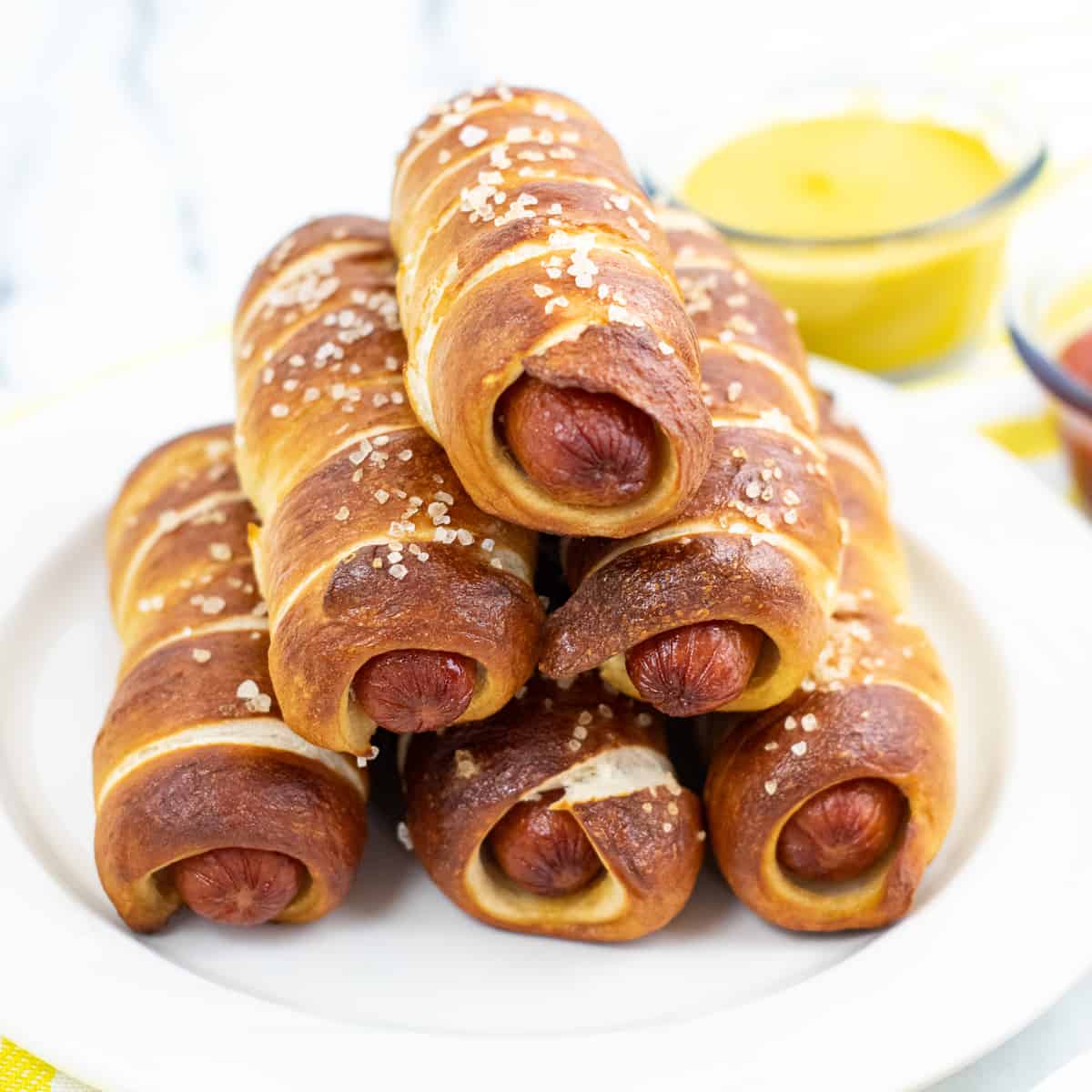 pretzel wrapped hot dogs stacked in a pyramid shape on white plate with bowl of mustard in the background.