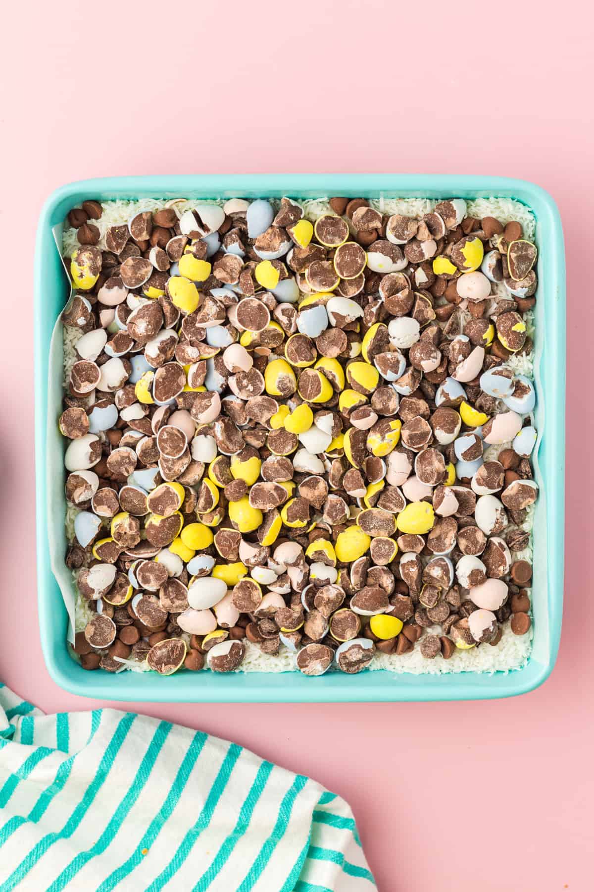 Crushed mini eggs on top of chocolate chips and shredded coconut in square baking pan.