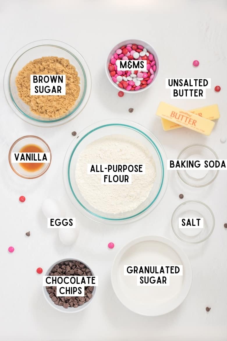 Two eggs, 2 sticks of butter, and bowls of: red, pink, and white M&Ms; light brown sugar; granulated sugar; salt; baking soda; chocolate chips; vanilla; and all-purpose flour