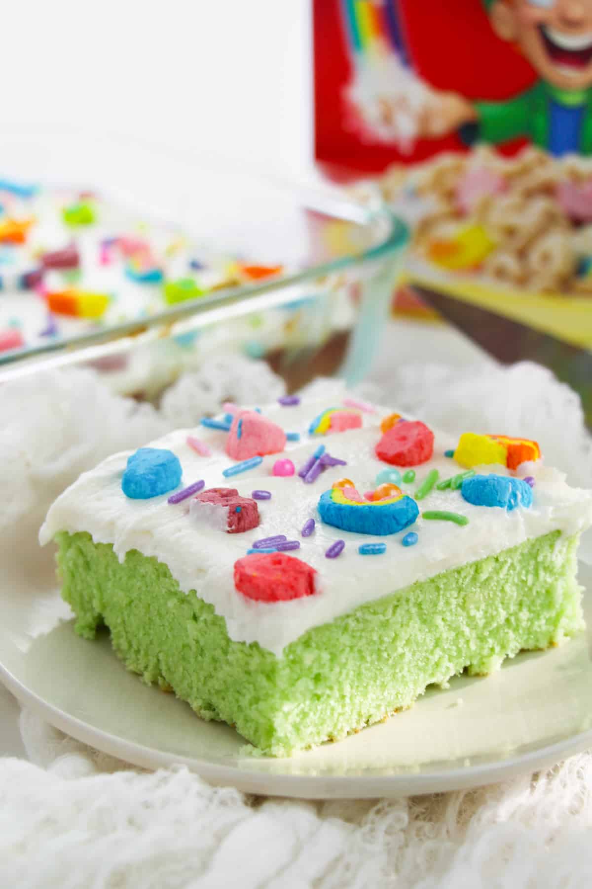 Green Lucky charms cake with white buttercream frosting, rainbow sprinkles, and lucky charms marshmallows on top. Box of Lucky Charms cereal is behind the slice of cake.