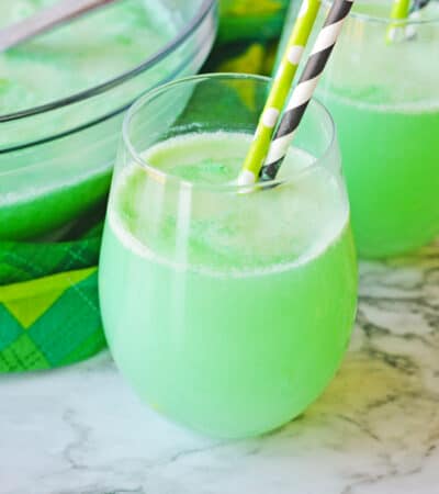 Shamrock Punch in a stemless wine glass with another glass of the green punch and punch bowl in the background.