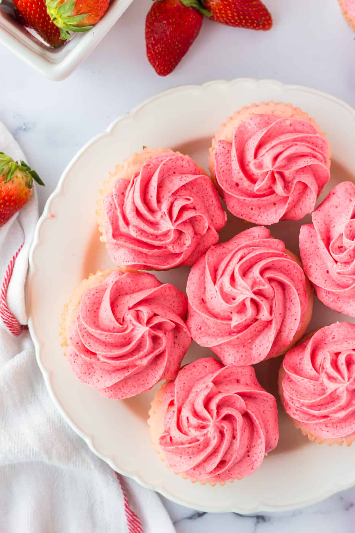 cupcakes with pink frosting arranged in a circle on white plate with strawberries in background