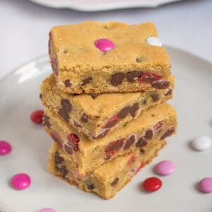 Four Valentine cookie bars with M&Ms stacked on top of one another on white plate with additional M&Ms sprinkled around them and more bars on bright pink plate in background.