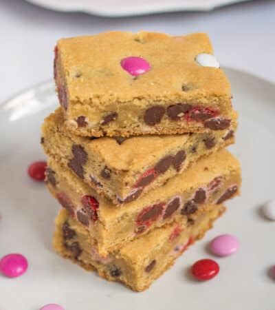 Four Valentine cookie bars with M&Ms stacked on top of one another on white plate with additional M&Ms sprinkled around them and more bars on bright pink plate in background.