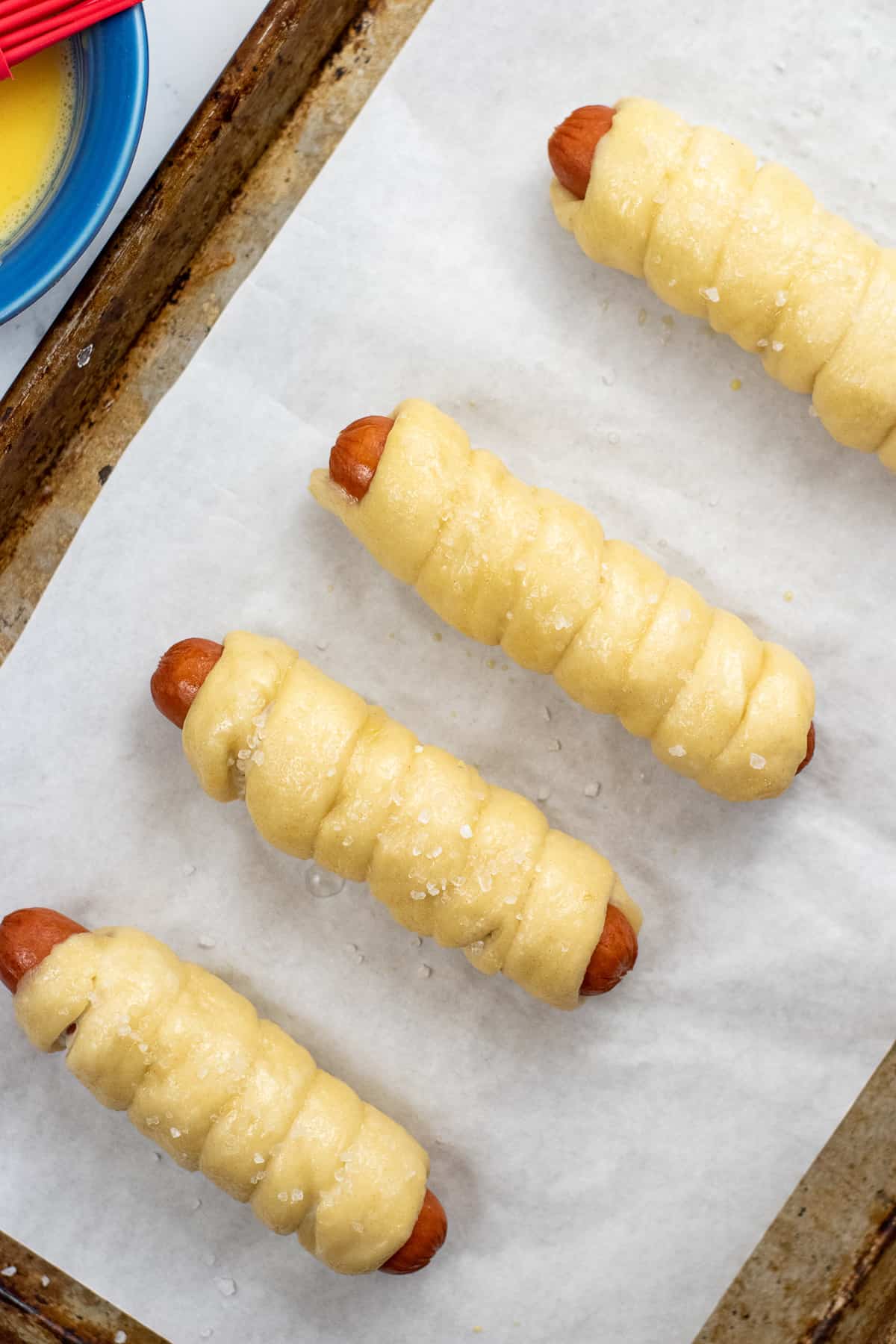 unbaked pretzel dough wrapped hot dogs with coarse salt on top on baking sheet.