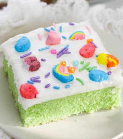 Slice of lucky charms cereal cake on white plate. The cake is bright green with white frosting topped with rainbow sprinkles and lucky charms marshmallows.