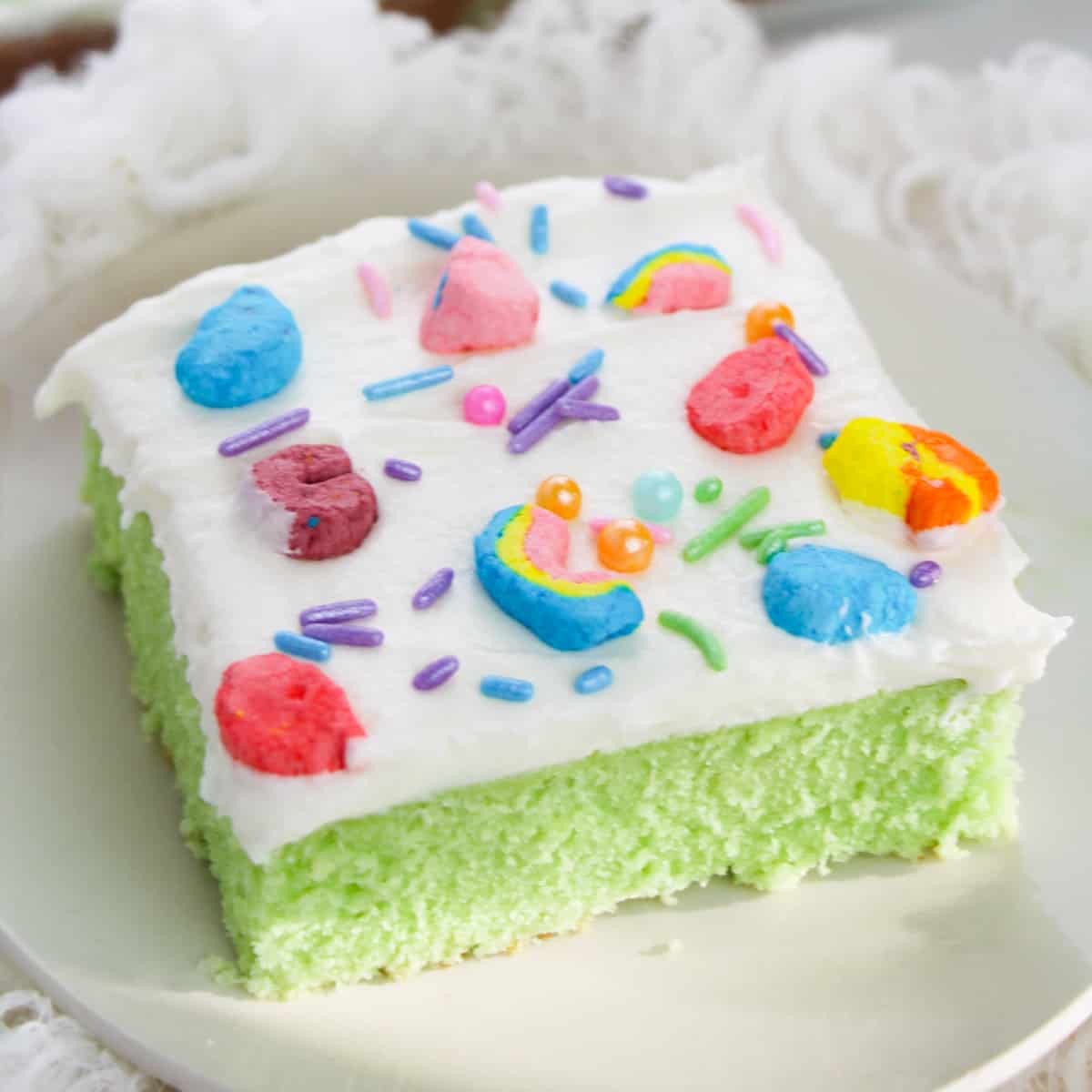 Slice of lucky charms cereal cake on white plate. The cake is bright green with white frosting topped with rainbow sprinkles and lucky charms marshmallows.