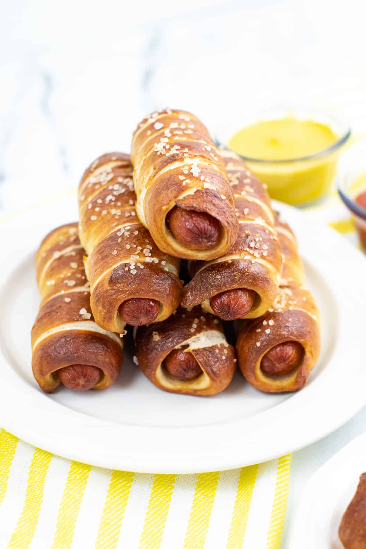 pretzel wrapped hot dogs stacked in a pyramid shape on white plate with bowl of mustard in the background.
