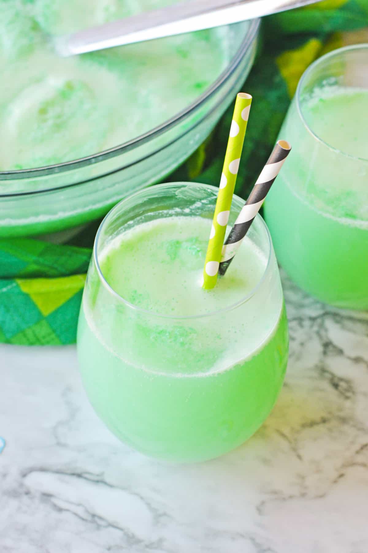 St. Patrick's Day Alcoholic Punch in stemless wine glass with two paper straws and punch bowl filled with green punch behind it.