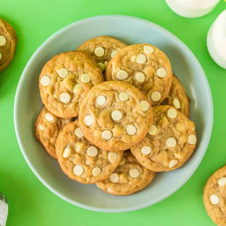 Banana pudding cookies with white chocolate chips on plate with glasses of milk and additional cookies in background.