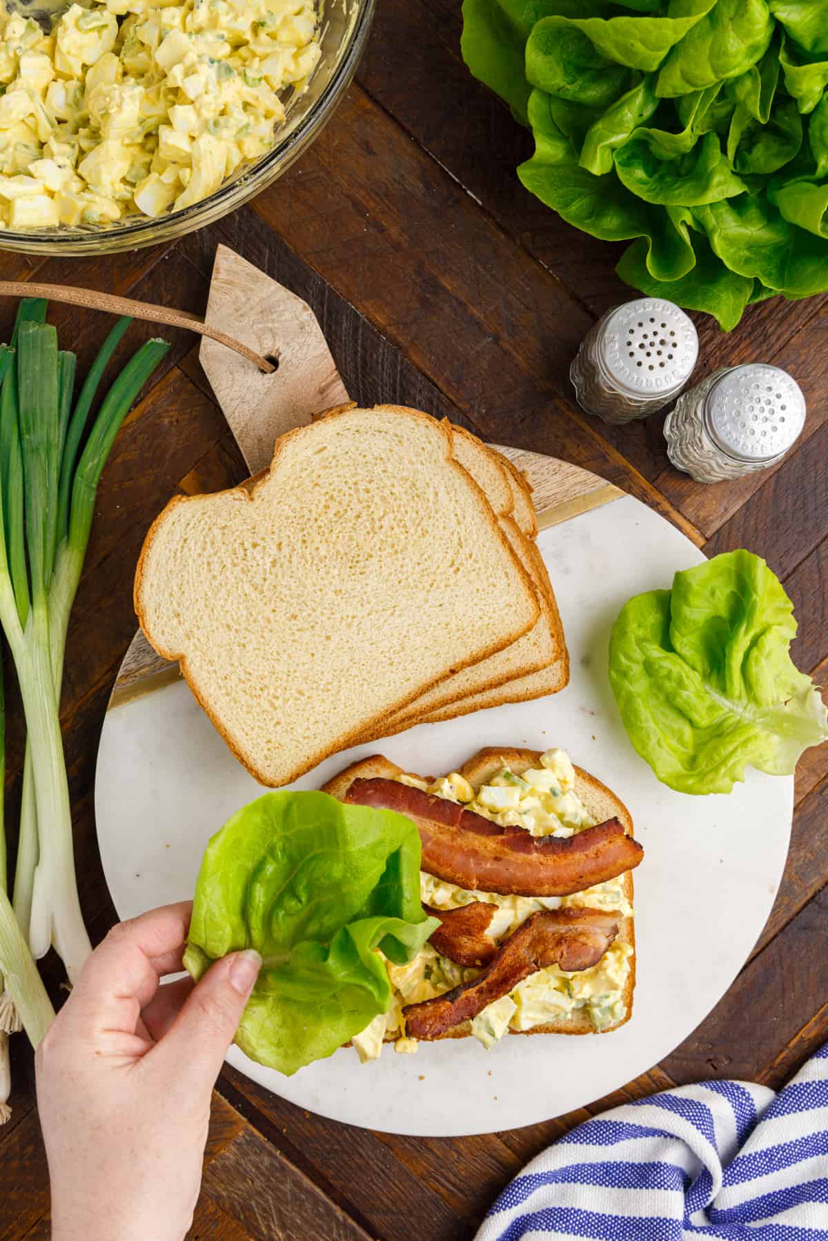 Hand placing lettuce on egg salad sandwich with bacon.