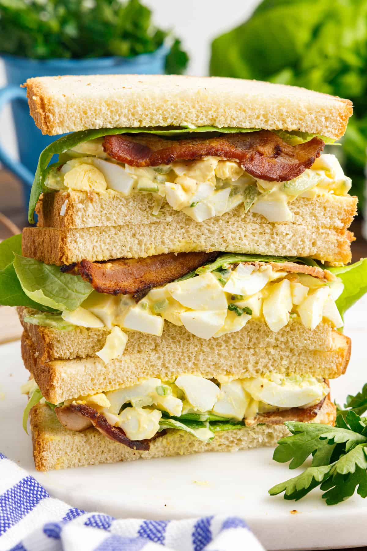 Thee halves of egg salad sandwiches with bacon and lettuce stacked on top of one another on a white plate.