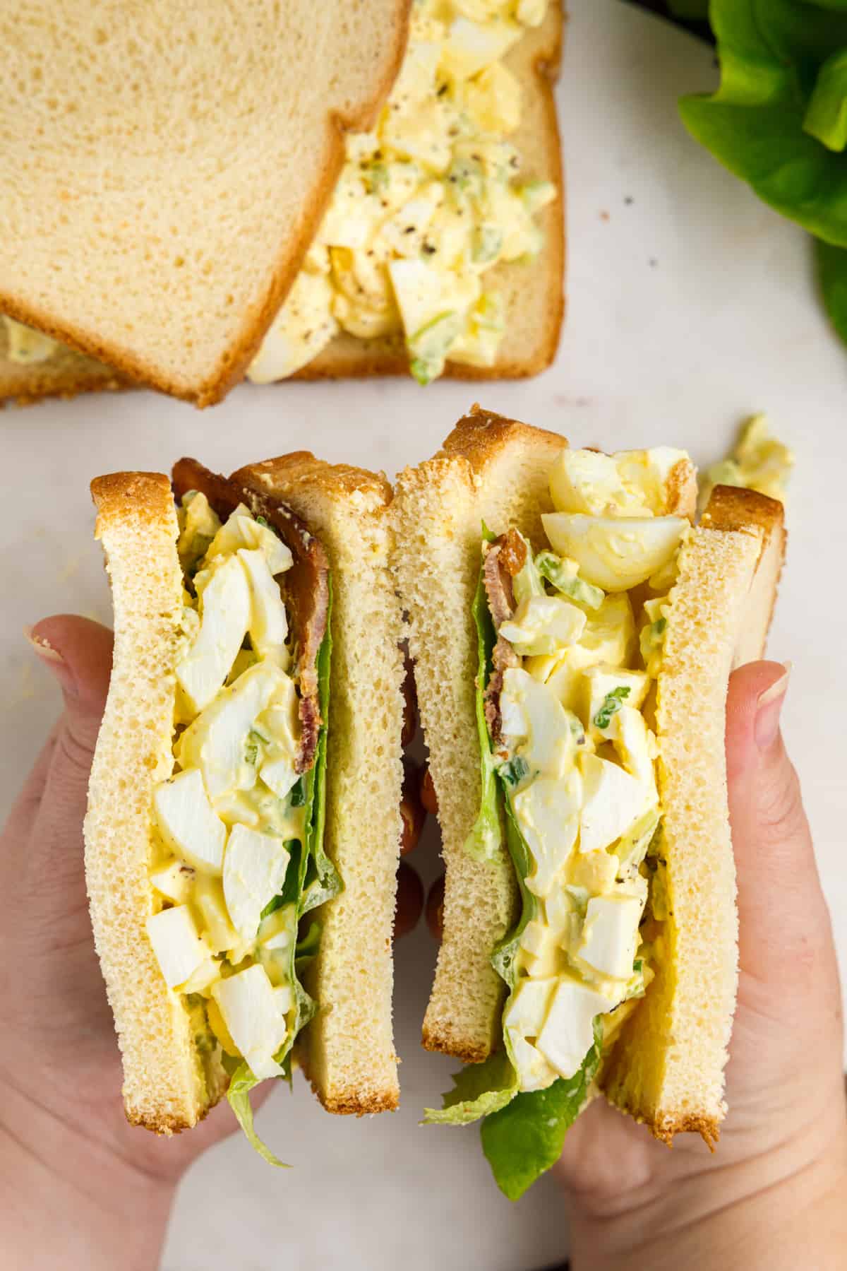 Two hands holding an egg salad sandwich with bacon and lettuce. An additional sandwich can be seen in the background.