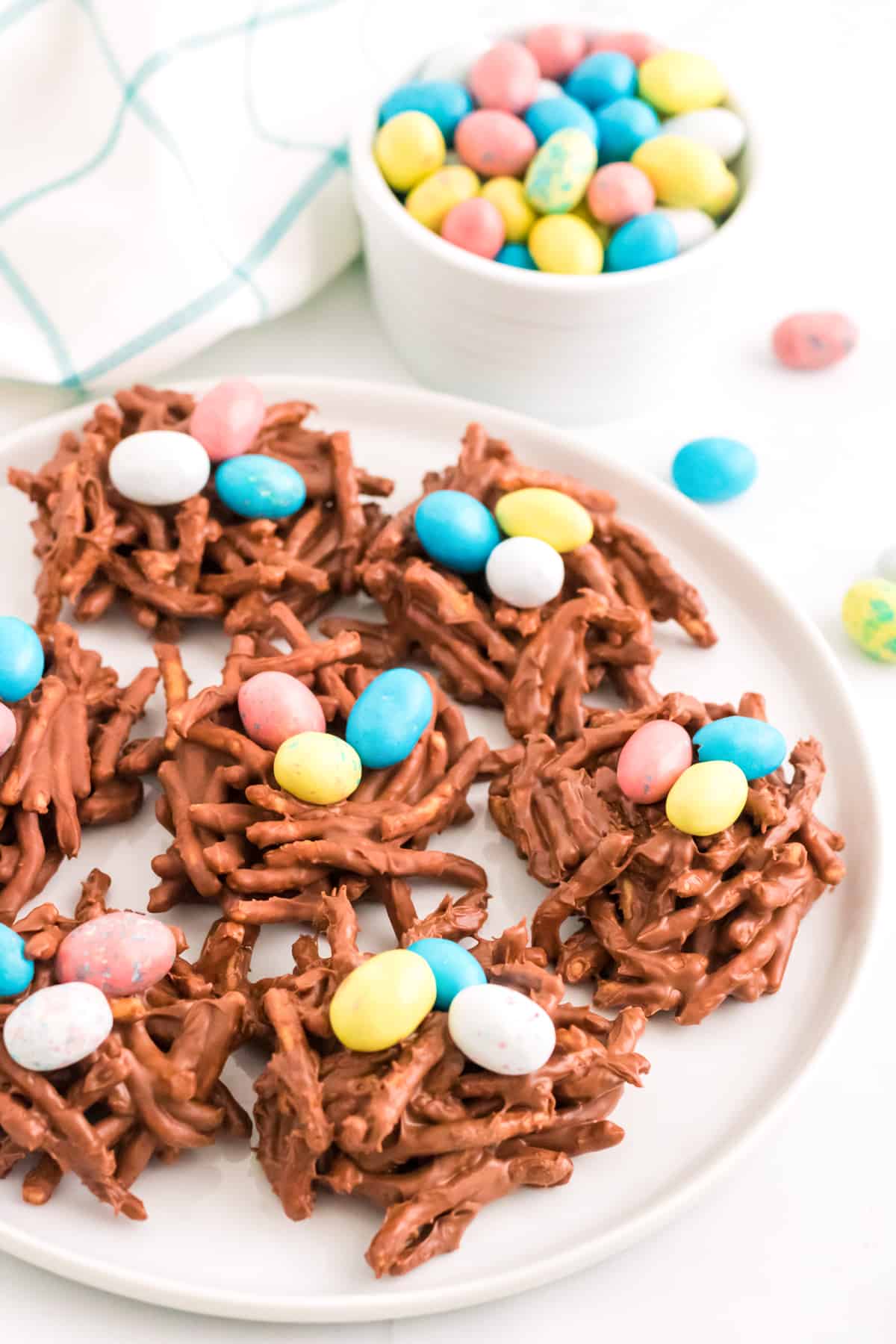 No-bake chocolate birds nest cookies arranged on a white plate with additional colored eggs in the background.