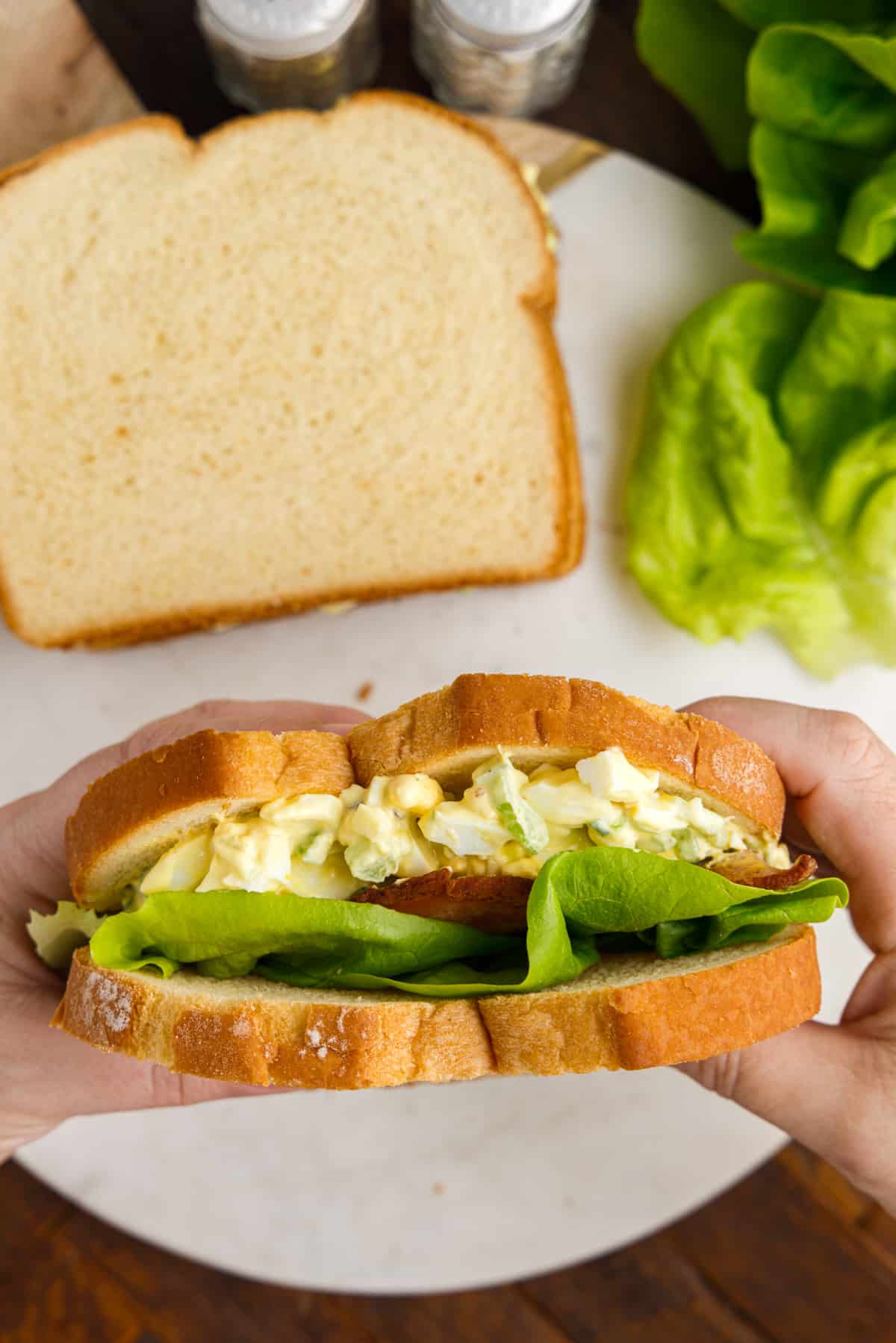 Hands holding an egg salad sandwich with lettuce and bacon on sandwich bread.