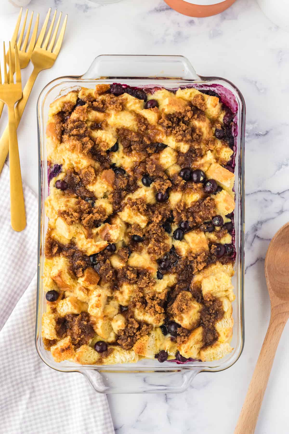Golden brown blueberry french toast casserole in baking dish with serving spoon and forks next to it.