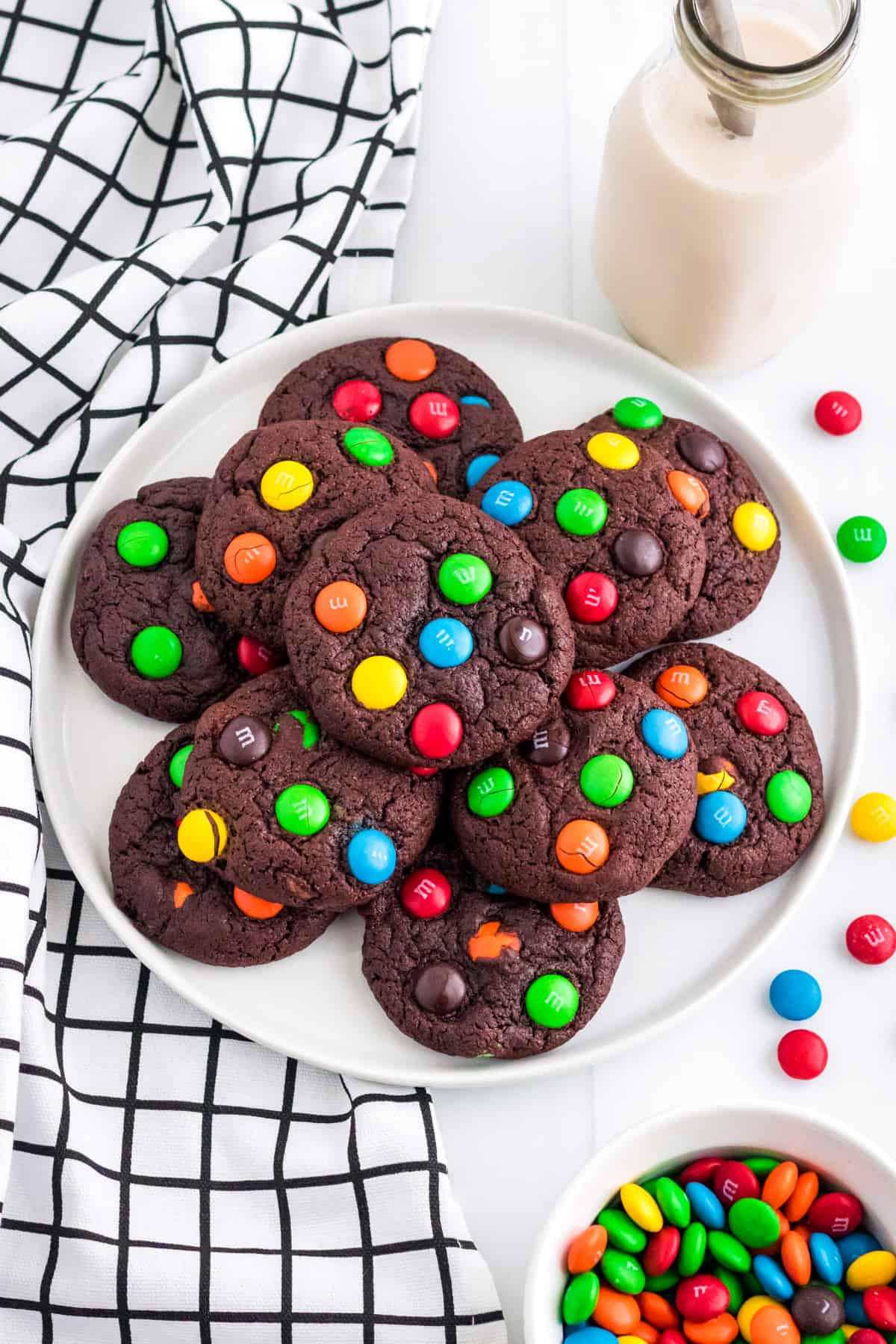 Chocolate cookies with M&Ms on white plate with glass of milk and more M&Ms next to them.