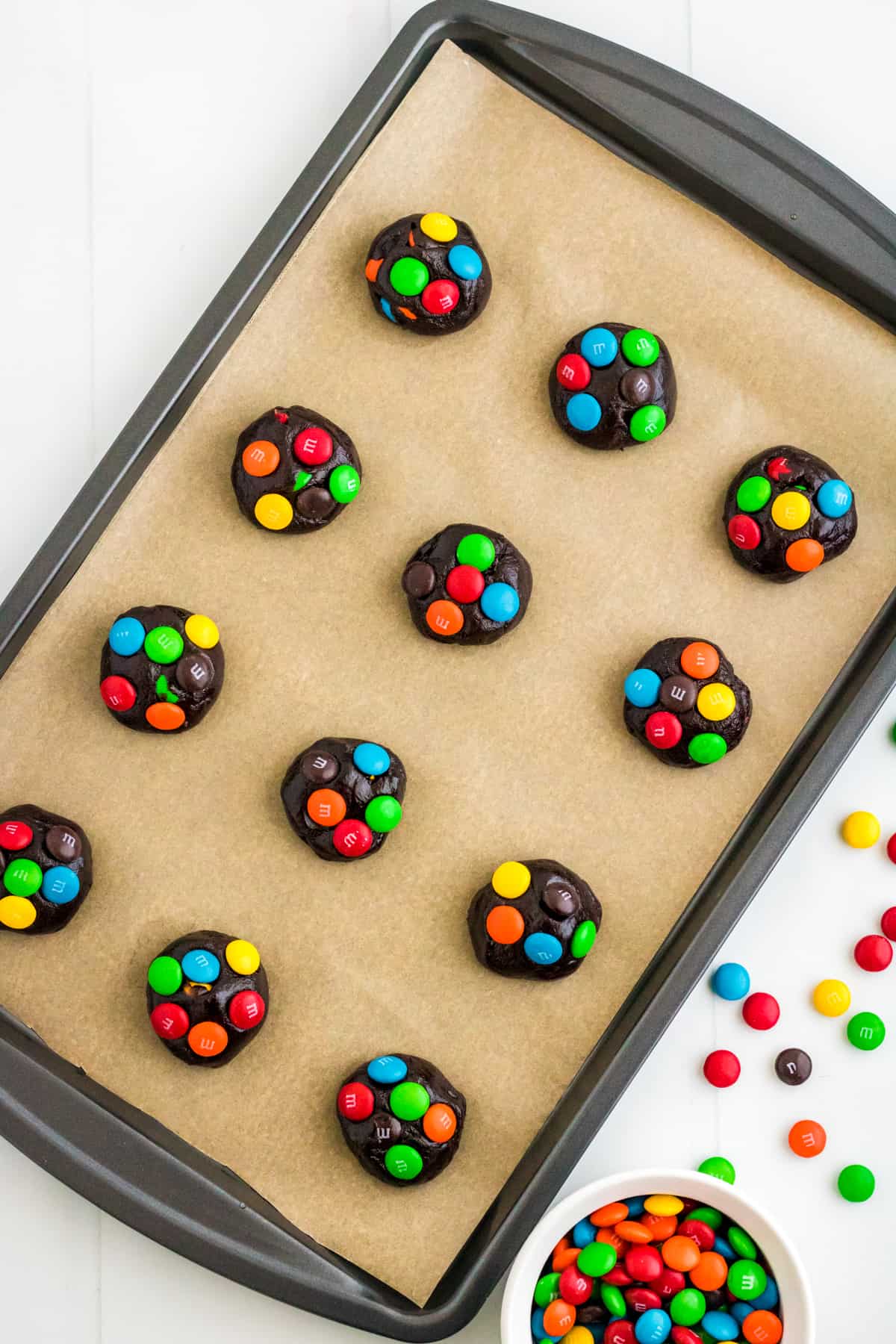 Balls of chocolate cake mix cookie dough with colorful M&Ms pressed into the top on a parchment lined baking sheet.