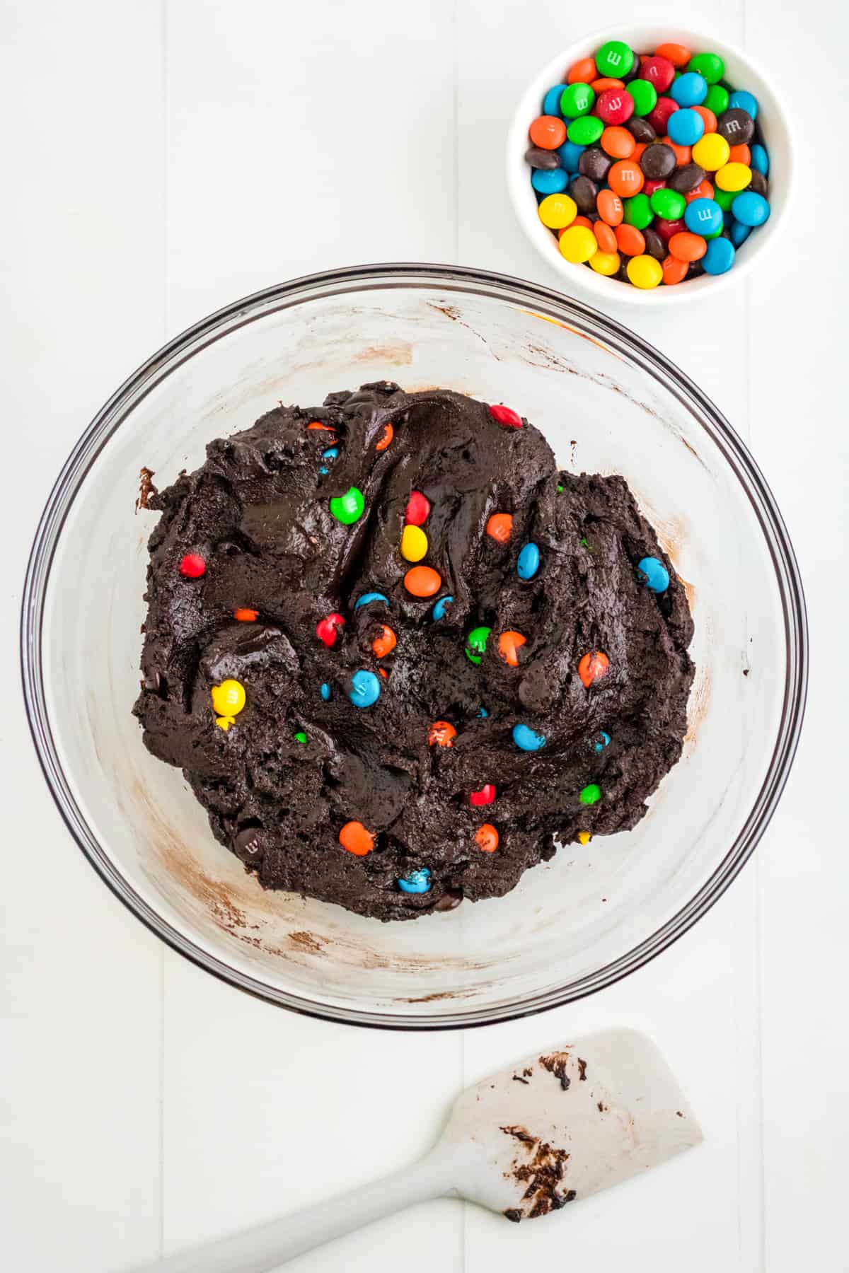 Chocolate M&M Cookie Batter in mixing bowl with spatula and M&Ms next to it.
