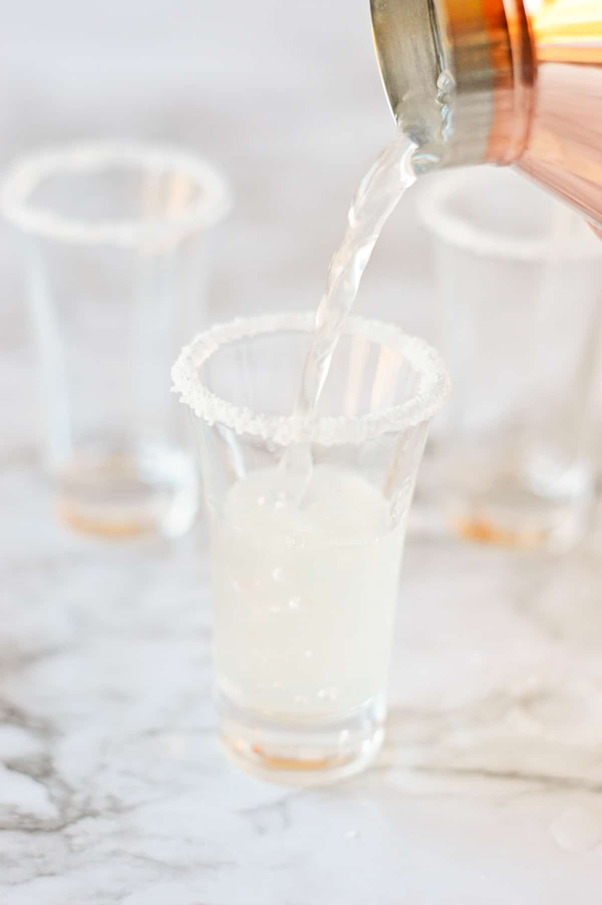 Cocktail shaker pouring lemon drop into rimmed shot glass.