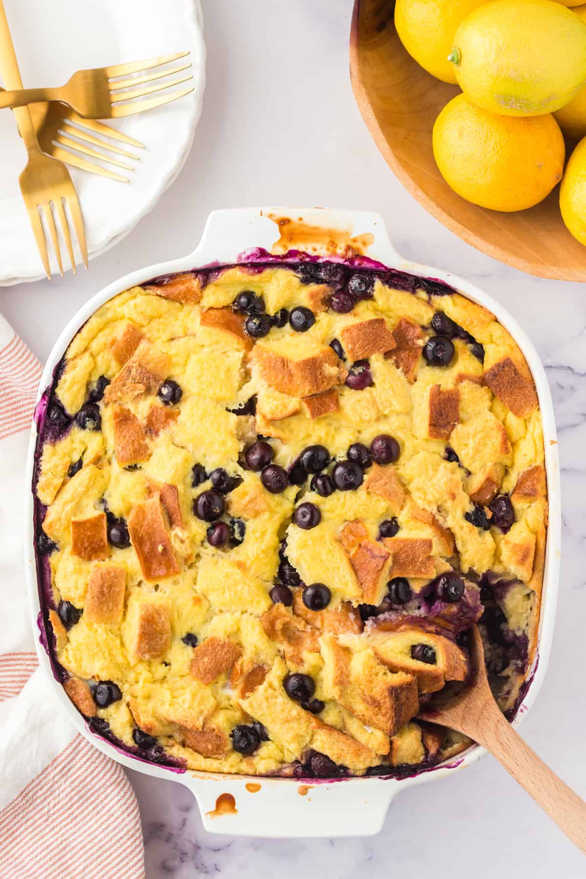 Lemon blueberry bread pudding in an square baking dish with wooden spoon sticking out of the corner.