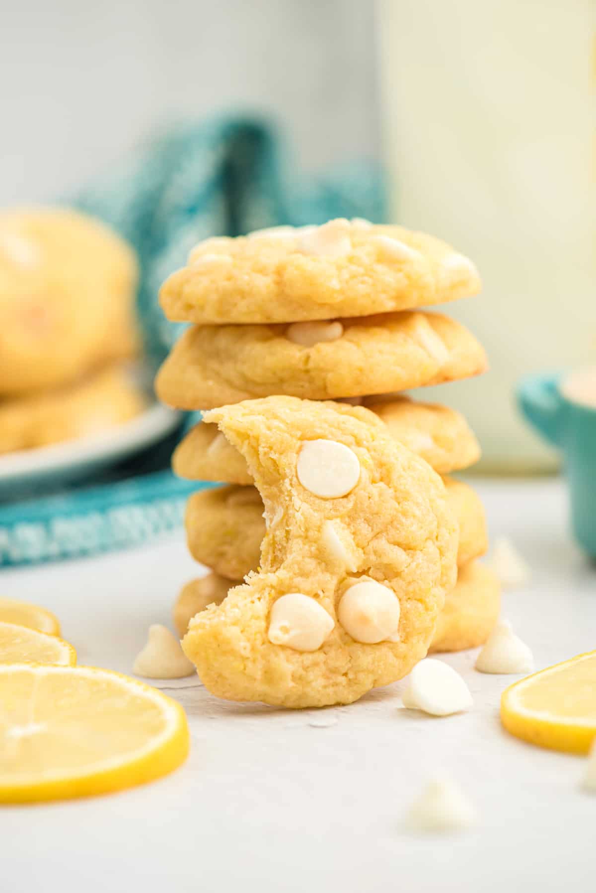 Lemon cookies with white chocolate chips stacked on top of one another. One cookie is leaning on front of the stack with a bite taken out of it.