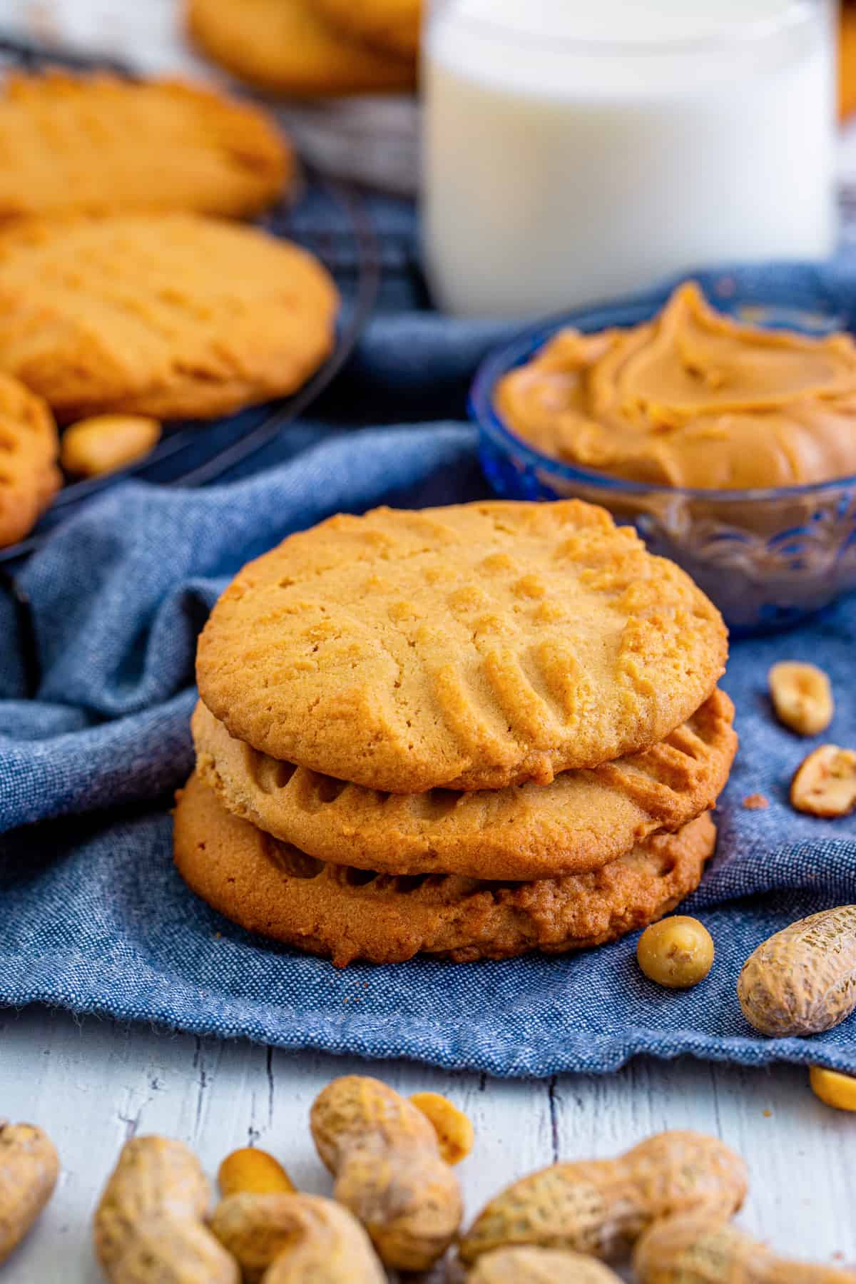 Three lunch lady peanut butter cookies stacked on top of one another with more cookies, peanut butter, peanuts, and glass of milk around it.