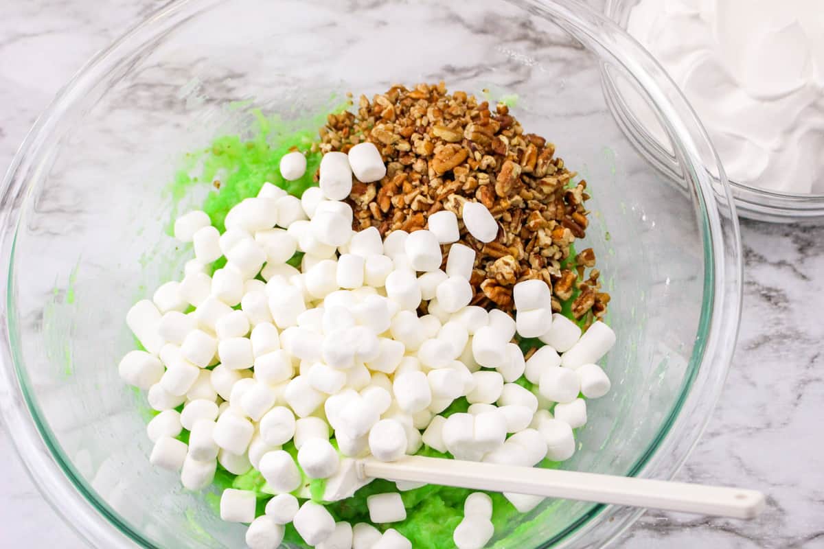 Mini marshmallows and chopped pecans on top of pistachio pudding mixture in mixing bowl.